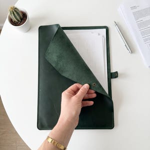 Puede incluir: Una carpeta de documentos de cuero verde oscuro con un cierre de solapa asegurado por un botón de presión de metal. La carpeta está abierta, revelando una pila de papel blanco en el interior. La superficie exterior tiene una textura mate.
