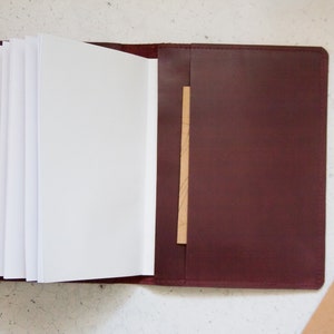 May include: A burgundy leather journal with a blank page open and a brown leather flap on the right side.