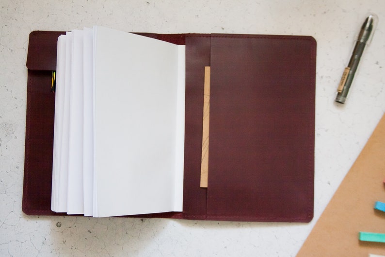 May include: A burgundy leather journal with a blank page open and a brown leather flap on the right side.