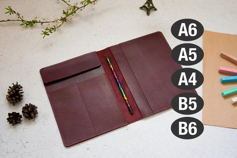 May include: Burgundy leather notebook cover with a pen holder and three pockets. The cover is open to show the inside. The cover is labeled with the sizes A6, A5, A4, B5, and B6.