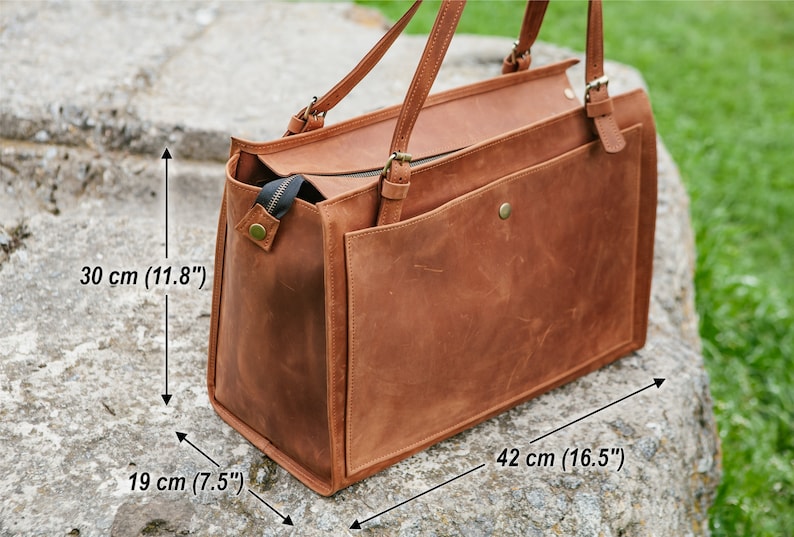 May include: A brown leather tote bag with a rectangular shape. The bag has two handles and a front pocket. Dimensions are 42 cm long, 30 cm high, and 19 cm wide. The bag has a zipper closure and brass-colored hardware.