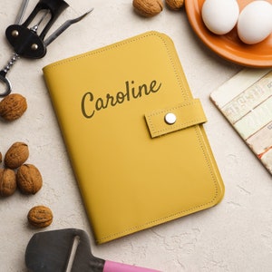 May include: A yellow leather journal with the name Caroline embossed on the front. The journal has a flap closure with a metal button.