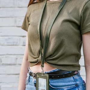 May include: A green leather lanyard with a green leather ID card holder. The lanyard is worn around the neck of a person wearing a green shirt and blue jeans.