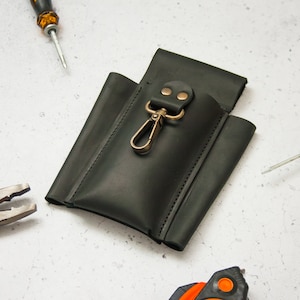 K&ouml;nnte beinhalten: Eine schwarze Ledertasche f&uuml;r Werkzeug mit einem Metallclip und zwei Messingnieten. Die Tasche liegt auf einer wei&szlig;en Oberfl&auml;che mit einem Schraubendreher und einer Zange daneben.