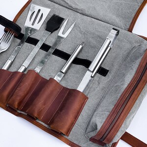 May include: A brown leather roll-up case with a gray canvas lining. The case contains six grilling tools: a spatula, a basting brush, a fork, a pair of tongs, a skewer, and a small tool.