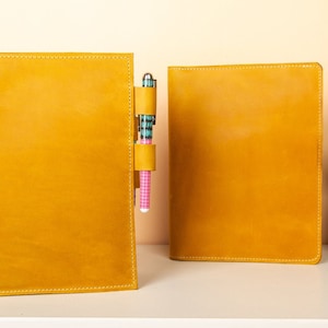May include: Two yellow leather-bound notebooks with a pen holder on the left notebook. The pen holder has a pink and blue patterned pen inside.
