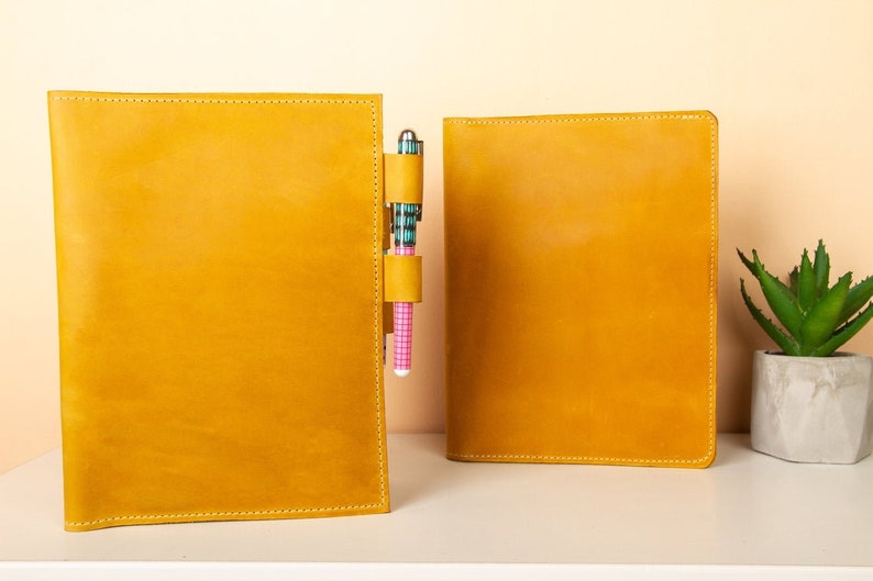 May include: Two yellow leather-bound notebooks with a pen holder on the left notebook. The pen holder has a pink and blue patterned pen inside.