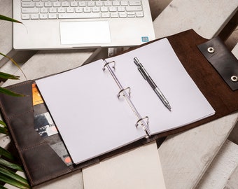 May include: A brown leather binder with a three-ring mechanism, open to blank white paper. A silver pen rests on the paper. The binder has card slots and a snap closure. A laptop is visible in the background.
