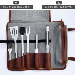 May include: A brown leather grilling tool roll with a gray canvas exterior. The roll is unrolled and shows six metal grilling tools, including a spatula, a basting brush, a fork, a knife, a pair of tongs, and a small tool. The roll has a zipper closure and a carrying handle. The roll is available in two sizes: M (50 x 41 cm or 19.7 x 16 inches) and L (57 x 50 cm or 22.5 x 19.5 inches).