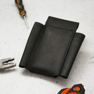 K&ouml;nnte beinhalten: Ein schwarzer Leder-Werkzeugg&uuml;rtel mit mehreren Taschen zum Verstauen von Werkzeugen. Der G&uuml;rtel liegt flach auf einer wei&szlig;en Oberfl&auml;che mit einer Zange, einem Schraubendreher und einem Ma&szlig;band in der N&auml;he.