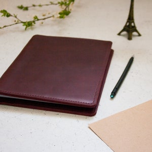 May include: A burgundy leather bound journal with a white page visible inside. The journal is on a white surface with a pencil, a small Eiffel Tower figurine, and a few pine cones.