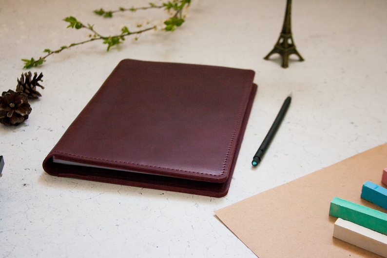May include: A burgundy leather bound journal with a white page visible inside. The journal is on a white surface with a pencil, a small Eiffel Tower figurine, and a few pine cones.