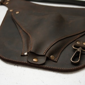 Pode incluir: Um cinto de couro castanho com uma bolsa grande anexada. A bolsa tem uma aba com fecho de press&atilde;o e um gancho de metal. O cinto &eacute; costurado com fio castanho.