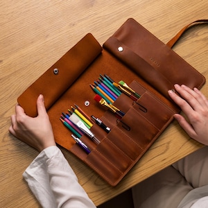 Puede incluir: Un estuche enrollable de cuero marrón, abierto para mostrar lápices de colores, pinceles y bolígrafos. El rollo está hecho de cuero marrón cálido y rico y tiene múltiples bolsillos. La palabra "BRONTE" es visible en la solapa. Para útiles de arte.