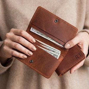 May include: A brown leather wallet with a matte finish, featuring a snap button closure and multiple interior slots containing paper currency. The exterior of the wallet is embossed with the capital letters RT in the bottom right corner.