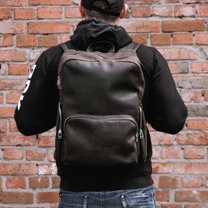 May include: A brown leather backpack with a zippered front pocket and adjustable straps. The backpack is being worn by a person standing in front of a brick wall.