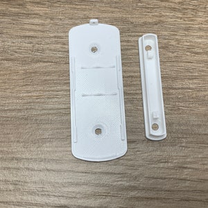 May include: Two white plastic wall mounts for a shelf or other object. The larger mount has two holes for screws and a slot for a shelf to slide into. The smaller mount has one hole for a screw and a slot for a shelf to slide into.