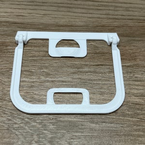 May include: A white 3D-printed plastic frame with a rectangular shape and rounded corners. It features a central circular cutout and a handle-like projection at the bottom. The frame has hinged sections on the top corners.
