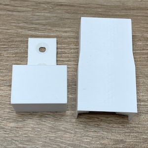 May include: Two white plastic pieces, one with a rectangular shape and the other with a more elongated shape. The rectangular piece has two small holes on the top.