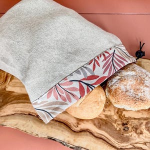 Op de afbeelding: Een beige linnen broodzak met een bloemenprint rand in roze en bruine tinten. De zak is gevuld met twee broden op een houten snijplank.