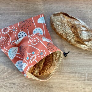 Peut inclure: Un sac en tissu réutilisable pour les produits avec un motif floral dans les tons orange, blanc et bleu. Le sac est en tissu et possède une fermeture à cordon. Une miche de pain est à l'intérieur du sac.