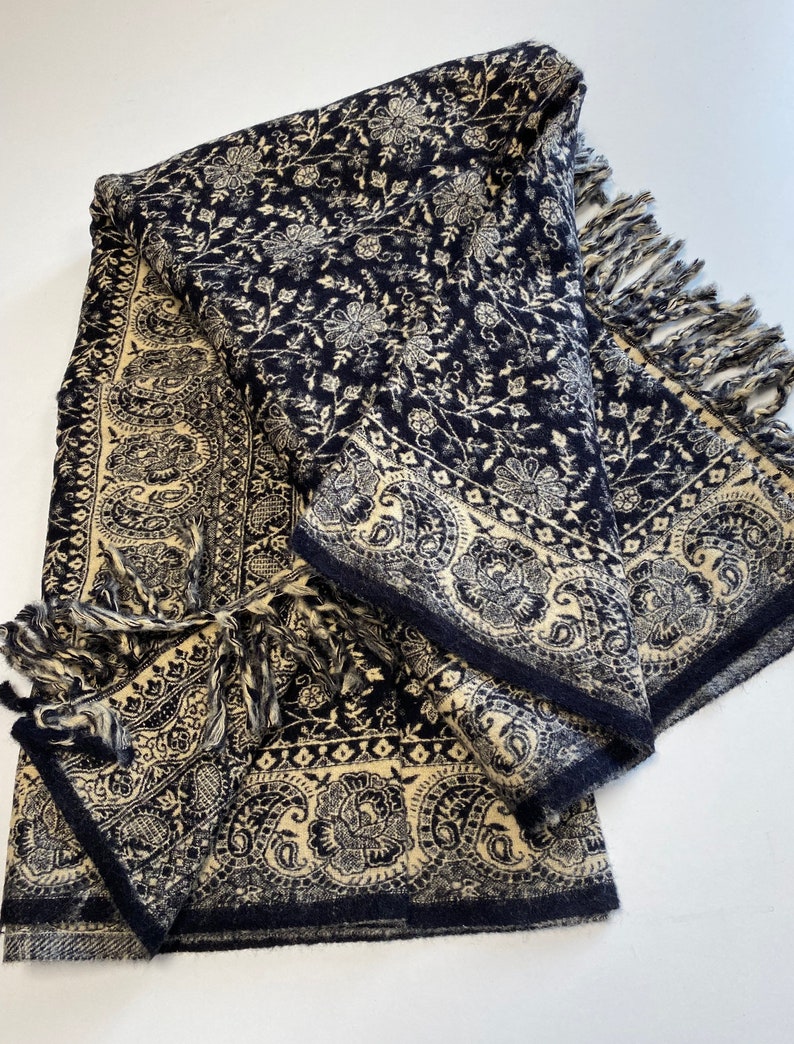 Puede incluir: Un chal plegado azul marino y crema con un estampado floral y cachemir. El chal tiene un borde con flecos y un dise&ntilde;o de borde detallado. La tela parece suave y texturizada, ideal para abrigo y estilo.