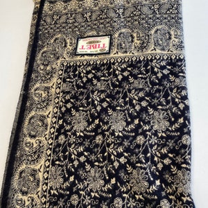 Puede incluir: Un chal de color azul marino y beige con un estampado floral y paisley. El chal tiene un borde con flecos y una etiqueta que dice "TIBET". El dise&ntilde;o presenta detalles intrincados y un borde contrastante.