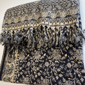Puede incluir: Una bufanda doblada de color azul marino y crema con un estampado floral y de cachemira. La bufanda tiene un borde con flecos y detalles intrincados. La bufanda est&aacute; hecha de un material suave.