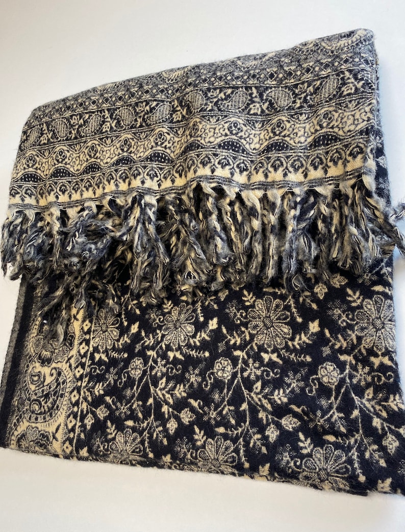 Puede incluir: Una bufanda doblada de color azul marino y crema con un estampado floral y de cachemira. La bufanda tiene un borde con flecos y detalles intrincados. La bufanda est&aacute; hecha de un material suave.
