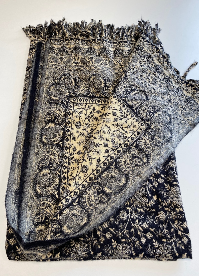 Puede incluir: Textil plegado con dise&ntilde;os florales y cachemir en azul marino y crema. La tela presenta patrones intrincados y un borde con flecos. Probablemente sea un chal, una manta o un pa&ntilde;o decorativo.