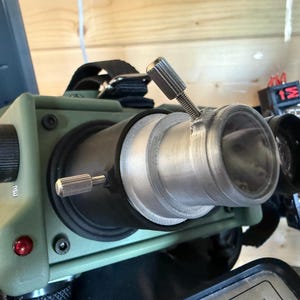 May include: A close-up of a military-style night vision device. The device is olive green with silver adjustment knobs and lenses. It has a black strap and various buttons and switches.