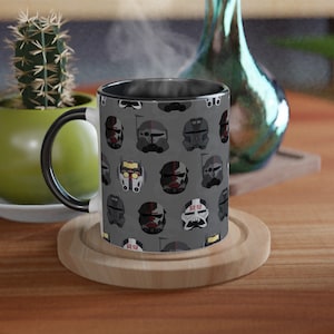 May include: A black and gray ceramic mug with a pattern of Star Wars clone trooper helmets. The mug has a black handle and is sitting on a wooden coaster.