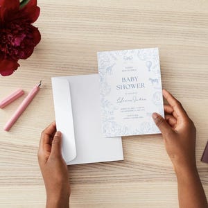 May include: A baby shower invitation with a blue floral and animal design. The invitation is held near a white envelope. A pink pen and a burgundy peony are on the wooden surface.