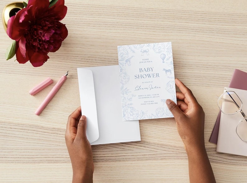 May include: A baby shower invitation with a blue floral and animal design. The invitation is held near a white envelope. A pink pen and a burgundy peony are on the wooden surface.