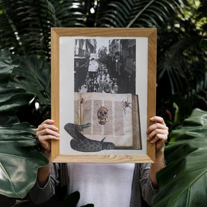 May include: A framed artwork featuring a black and white street scene and a collage of a skull, snake, and spiders over an open book. The artwork is held in front of a lush green foliage background.