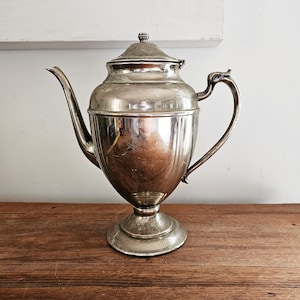 May include: A silver teapot with a rounded body and a fluted design. The teapot has a lid with a knob and a spout on the side. The teapot is sitting on a wooden surface.