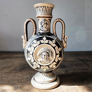 May include: A ceramic vase featuring a black and cream color scheme with two handles. The body displays a central circular relief of a person wearing a hat, surrounded by leaf motifs. A smaller relief of a person is on the neck, and the base is flared.