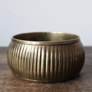 May include: A round, antique brass bowl with vertical ribbed detailing. The bowl has a wide, open top and a slightly rounded base. The brass has a warm, aged patina, suggesting it is vintage or antique. The bowl is likely used for decorative purposes.