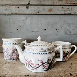May include: White ceramic tea set with a teapot, two mugs, and a creamer. The set features a floral design with pink blossoms and black branches. Blue dots and decorative trim accent the rims and lids. The set is placed on a wooden surface.