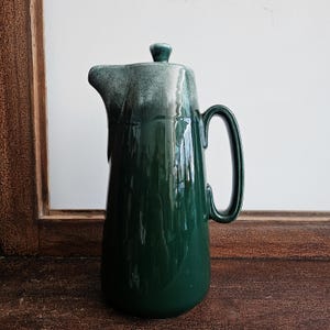 May include: A tall, glossy green ceramic pitcher with a matching lid. The pitcher has a curved handle and a spout for pouring. The top of the pitcher and lid have a lighter, faded green color. The pitcher is likely used for serving beverages.