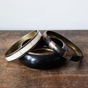 May include: A collection of four bangles in various designs. One bangle has a brass finish with white and black rectangular inlays. Another is black with a brass finish. The third has a black, copper, and silver design. The fourth has a brass finish.