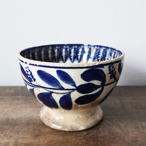 May include: A small, off-white ceramic bowl with a pedestal base. The bowl features a blue leaf and floral pattern around the exterior and a blue patterned rim. The bowl is likely used for serving small portions of food.