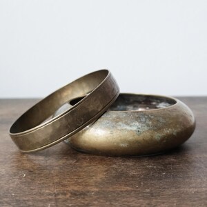 May include: Two antique brass bowls on a wooden surface. One is a shallow, round bowl, and the other is a matching ring-shaped bowl. The bowls have a hammered texture and a tarnished patina, giving them a vintage look.