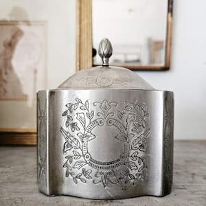May include: A silver-colored, antique, lidded box with ornate floral and leaf engravings. The box has a geometric shape with a decorative finial on the lid. The design includes a central circular emblem surrounded by detailed botanical patterns.