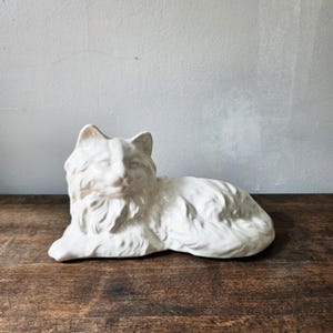 May include: A white ceramic figurine of a long-haired cat in a reclining position. The sculpture features detailed molded fur textures, pointed ears, and a smooth, glossy finish, capturing the feline in a resting pose with its head turned forward.