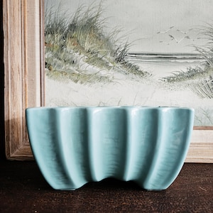 May include: A light blue ceramic planter with a ribbed design. The rectangular planter has a curved base and is set against a vintage painting of a beach scene.