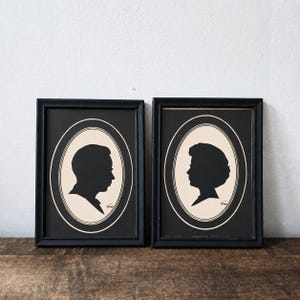 May include: Two rectangular black frames containing oval-shaped silhouettes of a person in profile. Each silhouette is rendered in black against a cream-colored background, enclosed within a thin black oval border. Both portraits feature a small signature.