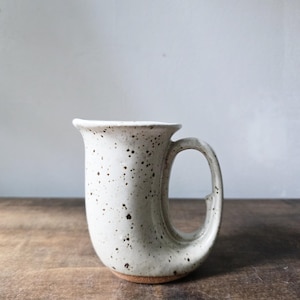 May include: A ceramic mug with a speckled off-white glaze and a curved, horn-like silhouette. The vessel features a wide, flared rim and a large, integrated handle that extends from the top to the base, revealing a natural brown clay bottom.