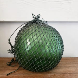 May include: A spherical green glass fishing float encased in a dark grey knotted rope net. The glass surface is smooth and translucent, showing light reflections, while the surrounding netting features a consistent diamond-shaped pattern.
