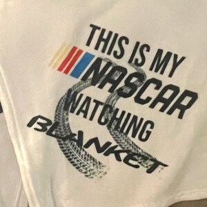 May include: Two white fleece blankets with the text "This is my NASCAR watching blanket" printed on them. The blankets feature a tyre tread design and a red, white and blue chequered flag.